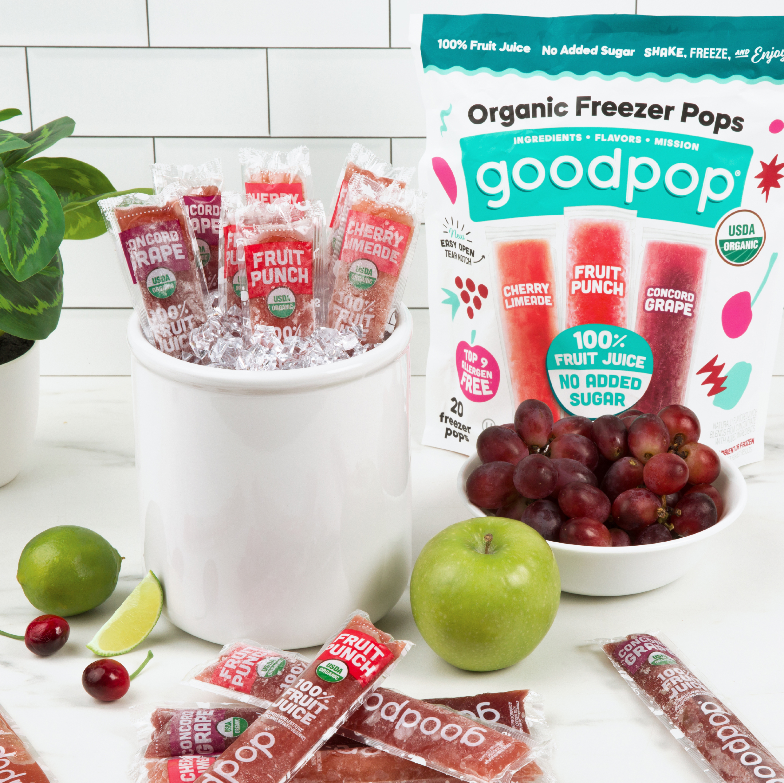 Crock of ice sits filled with ice and GoodPop Organic Freezer Pops sits on a kitchen counter surrounded by more pops and fresh apples, cherries, grapes and limes